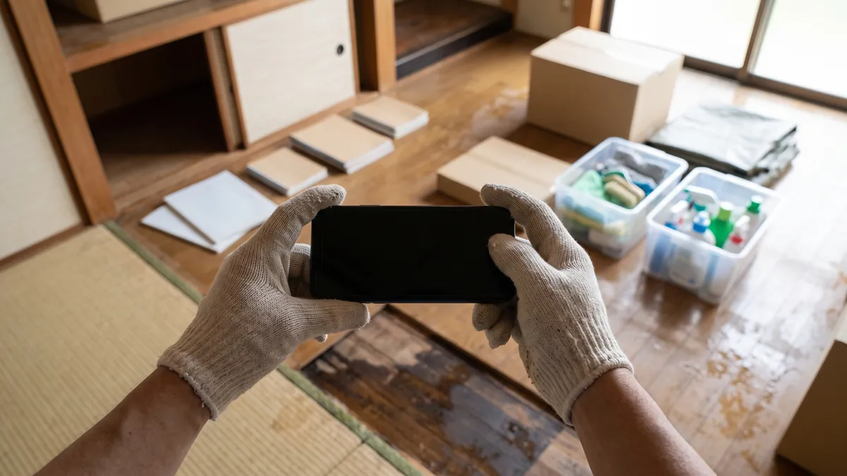片付け前の室内を記録するため、スマートフォンを構えた両手の手元と、その先の濡れた床と家具
